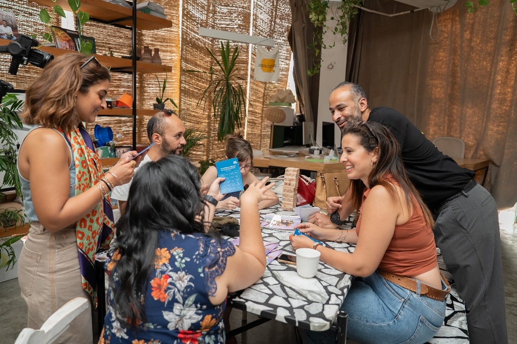 A group around a table in a plant-filled studio, mid-conversation over cards and a game